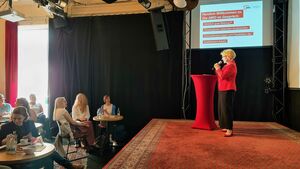 Man sieht Katja Glybowskaja seitlich auf einer Bühne mit einem Mikrofon in der Hand an einem roten Stehtisch stehend. Sie sprich zu einem kleinen Publikum, was vor ihr an runden Tischen sitzt. 