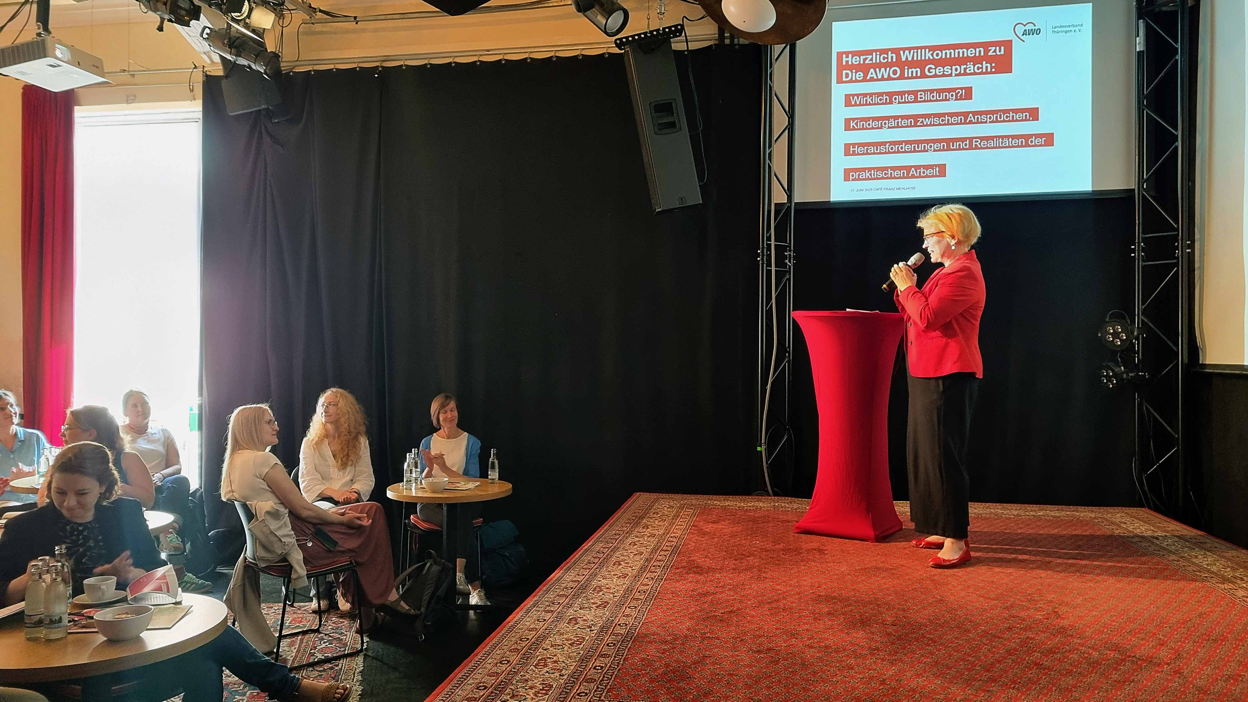 Man sieht Katja Glybowskaja seitlich auf einer Bühne mit einem Mikrofon in der Hand an einem roten Stehtisch stehend. Sie sprich zu einem kleinen Publikum, was vor ihr an runden Tischen sitzt. 